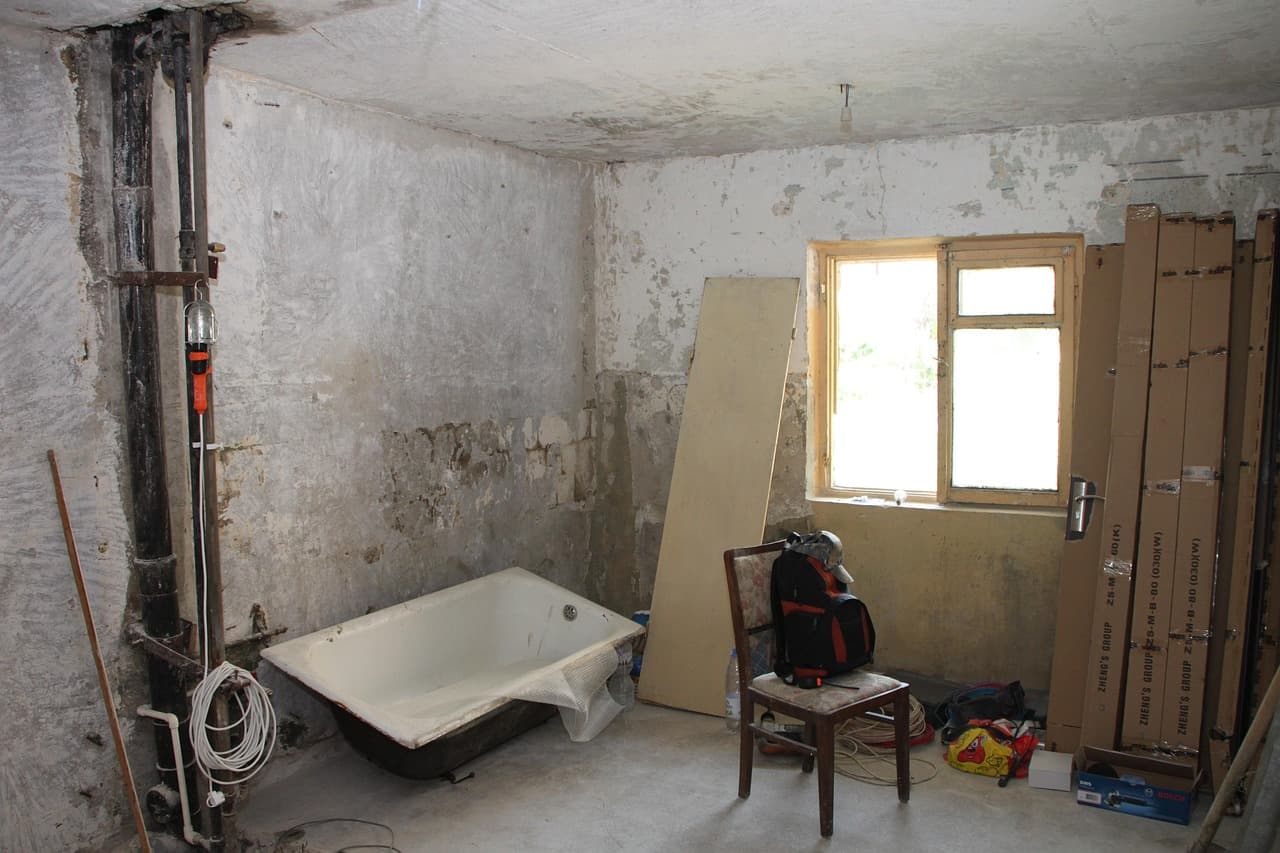 A bathroom stripped back to the walls mid-refit, the old bath still in place and boards stacked by the window